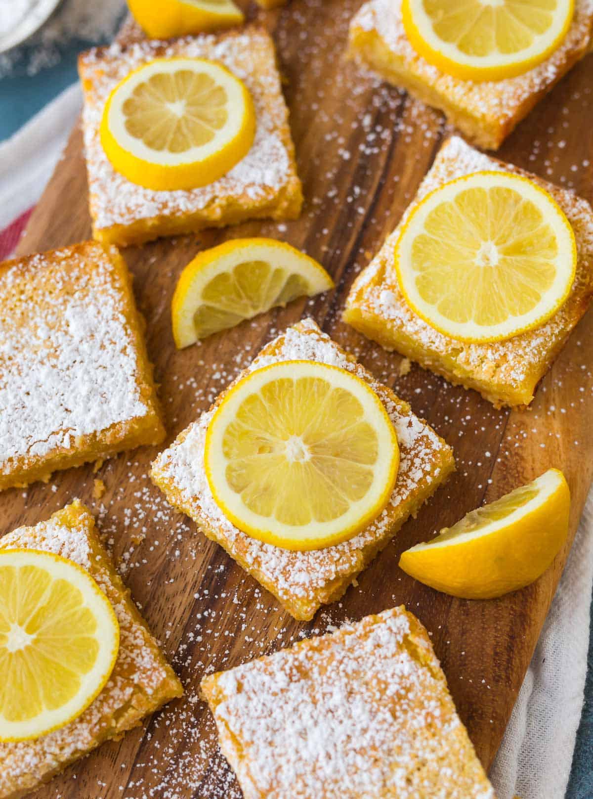 Close-up photo of lemon bars cut into squares on a butcher block board with powdered sugar and lemon slices