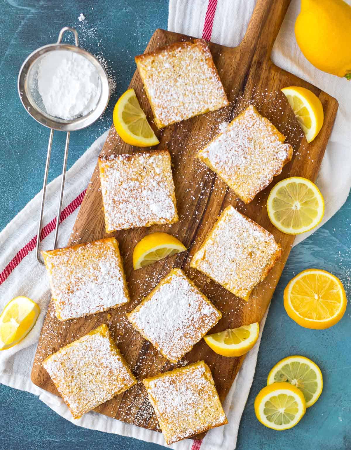 Homemade lemon bars dusted with powdered sugar on a wood serving board with lemon slices and wedges
