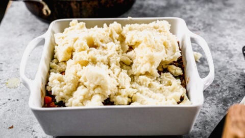 Mashed potatoes being added on top of ingredients in a baking dish