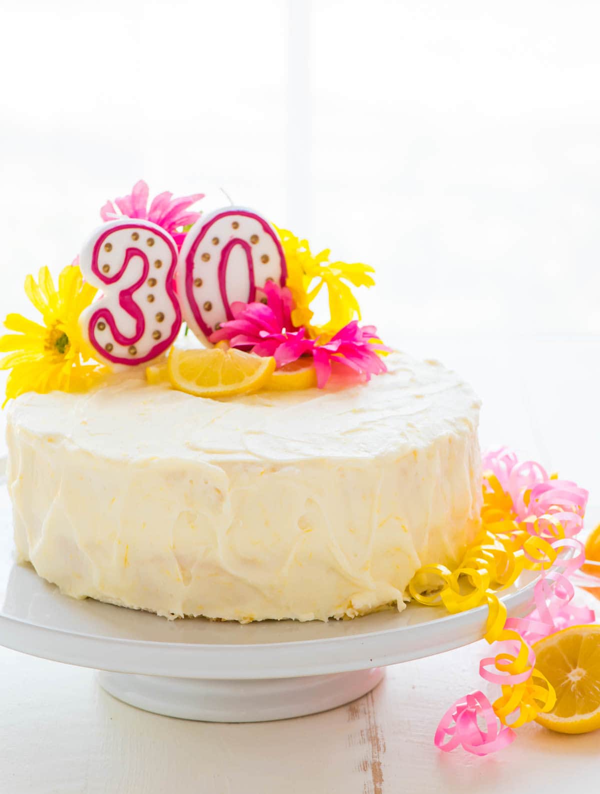 Lemon layer cake with lemon cream cheese frosting on a cake stand with birthday candles, ribbons and fresh flowers
