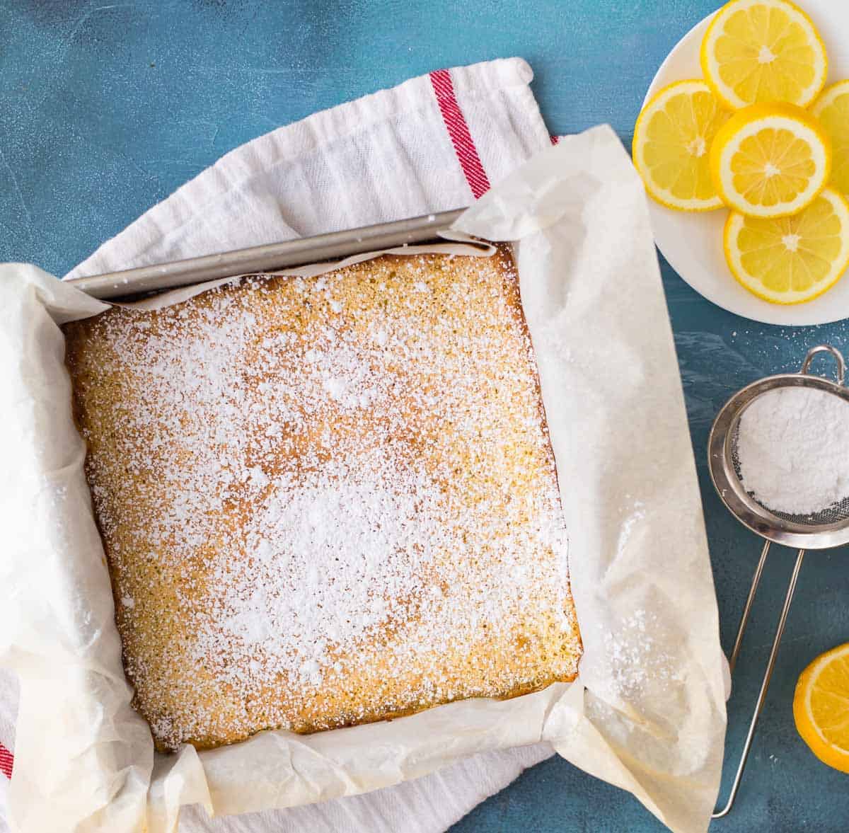The best lemon bars baked in a square pan dusted with powdered sugar on a blue background