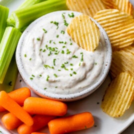 A white bowl of Greek yogurt ranch dip with carrots and celery on the side and a chip in the dip