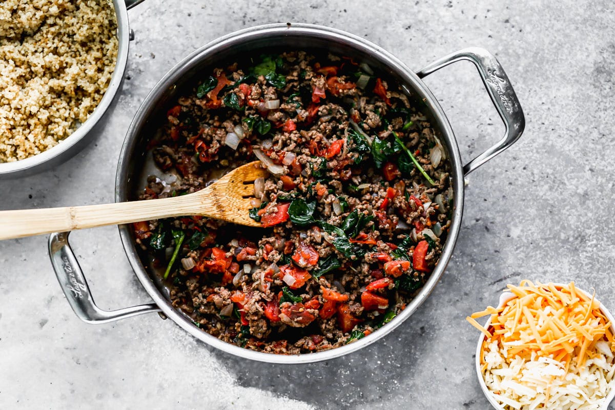 A skillet with ground meat and vegetables with bowls of quinoa and shredded cheese next to it